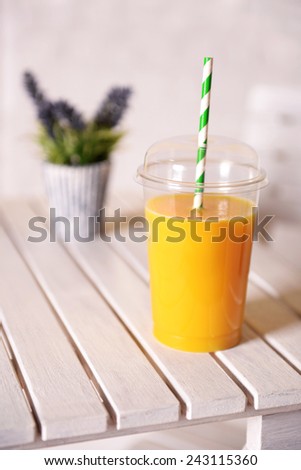 Orange juice in fast food closed cup with tube on wooden table with plant and light wall background
