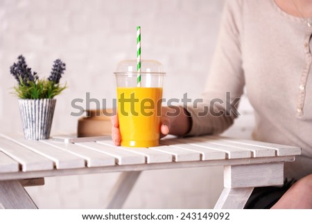 Female hand at wooden table with fast food closed cup of orange juice and near books and plant on light wall background