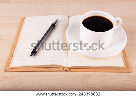 Cup of coffee on saucer with open notebook and pen on wooden table background