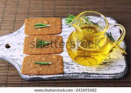 Crispbread with sprigs of rosemary on wooden cutting board with jug of oil on bamboo mat background