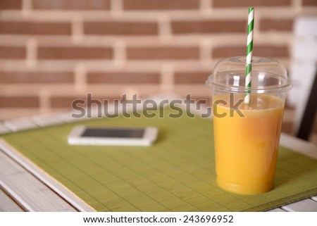 Orange juice in fast food closed cup with tube and mobile phone on wooden table and brick wall background