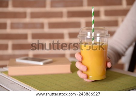 Female hand at table with fast food closed cup of orange juice on brick wall background