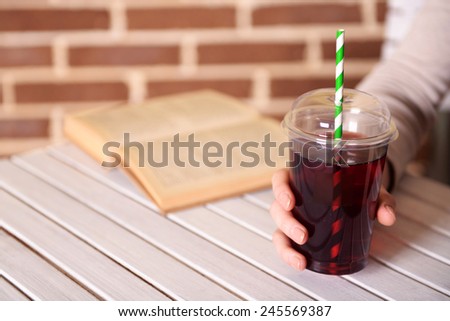 Female sitting at wooden table with fast food closed cup of pomegranate juice and near book on brick wall background