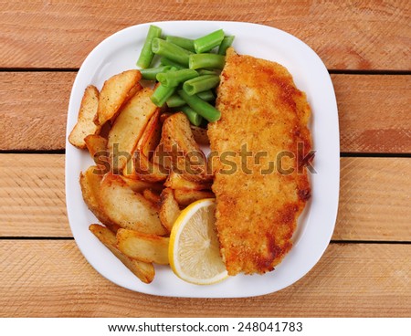 Breaded fried fish fillet and potatoes with asparagus and sliced lemon on plate and wooden planks background