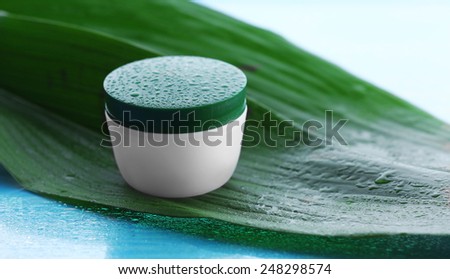 Cosmetic cream with water drops on green leaves on light background