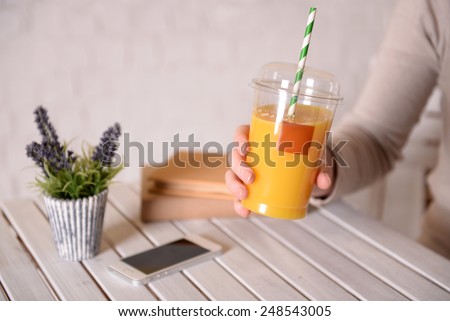 Female hand at wooden table with fast food closed cup of orange juice and near books, plant and mobile phone on light wall background