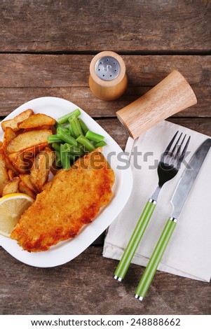 Breaded fried fish fillet and potatoes with asparagus and lemon on plate and wooden planks background