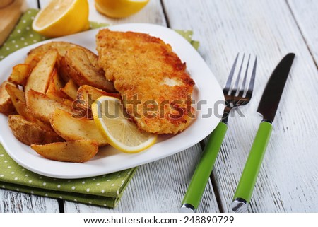 Breaded fried fish fillet and potatoes with sliced lemon on plate with napkin on color wooden planks background