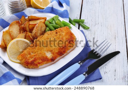Breaded fried fish fillet and potatoes with asparagus and sliced lemon on plate with napkin on color wooden planks background