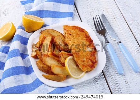 Breaded fried fish fillet and potatoes with sliced lemon on plate with napkin on color wooden planks background
