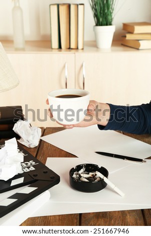 Female hand with cup of coffee at desktop with moving clapper and working mess on light cupboard background