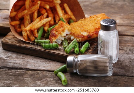 Breaded fried fish fillet and potatoes in paper bag with asparagus on cutting board and rustic wooden background