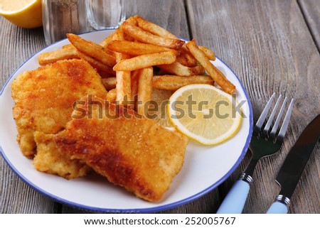Breaded fried fish fillets and potatoes with with sliced lemon and cutlery on plate and wooden planks background