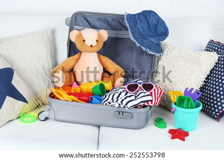 Suitcase packed with clothes and child toys on sofa with pillows and white brick wall background