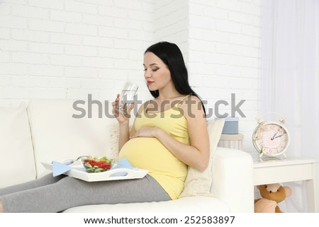 Young pregnant woman relaxing on sofa with glass of water and salad on home interior background