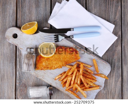 Breaded fried fish fillet and potatoes with sliced lemon and cutlery on cutting board and wooden planks background