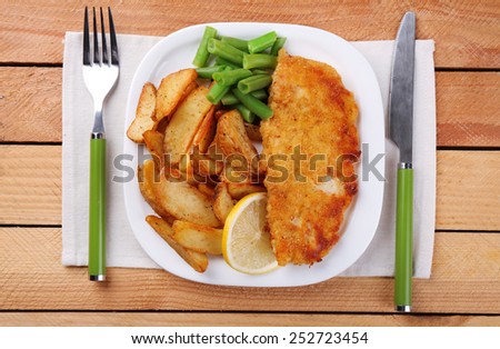 Breaded fried fish fillet and potatoes with asparagus and lemon on plate with napkin and cutlery on wooden planks background
