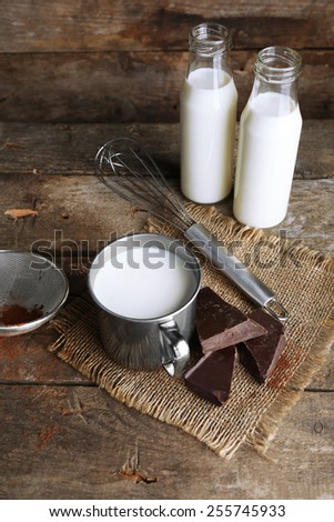 Metal mug and glass bottles of milk with chocolate chunks and strainer of cocoa on burlap cloth and rustic wooden planks background