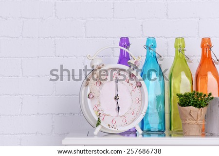 Interior design with alarm clock, plant and decorative colorful glass bottles on tabletop on white brick wall background