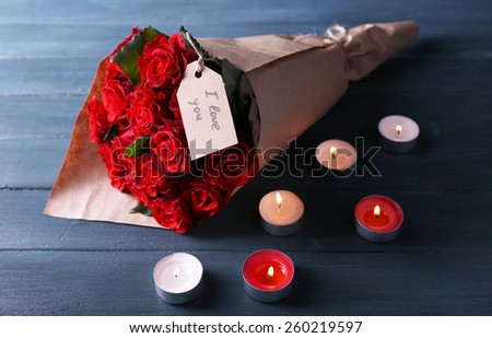 Bouquet of red roses with tag wrapped in paper and candles on wooden background