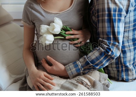 Young pregnant woman sitting with husband and holding flowers in room