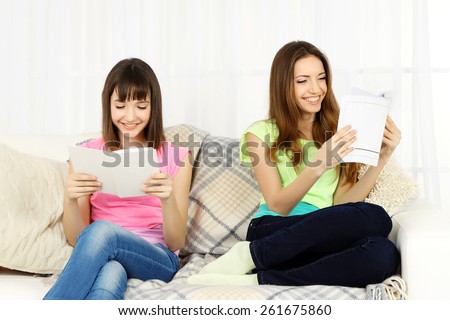 Two girls sitting on sofa and reading book on home interior background