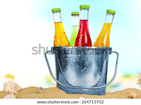 Bottles of tasty drink in metal bucket with ice on sand on bright background