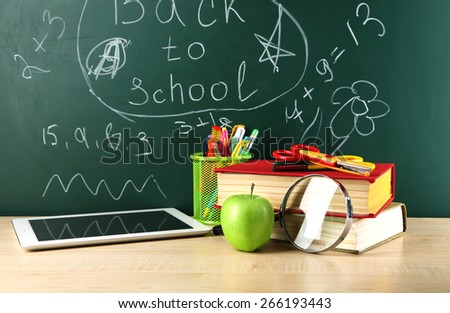 Digital tablet, books, colorful pens and apple on desk in front of blackboard