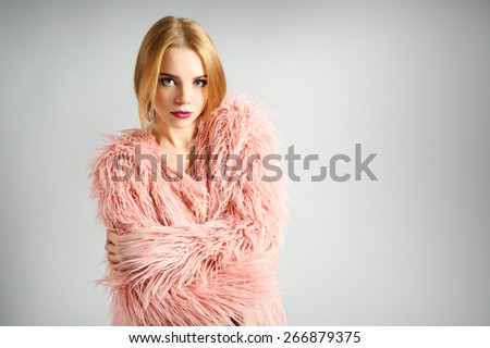 Expressive young model in pink fur coat and black dress on gray background