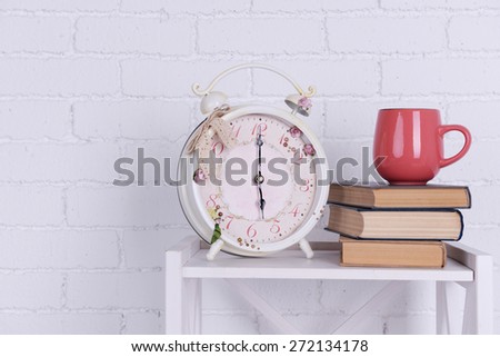 Interior design with alarm clock, stack of books and pink cup on tabletop on white brick wall background