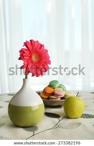Color gerbera flower in vase and apple with macaroons on table on curtains background