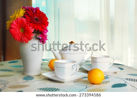 Colorful gerbera in vase with teapot, cups and lemons on table on curtains background