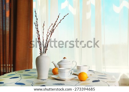Willow twigs in vase with teapot, cups and lemon on table on curtains background