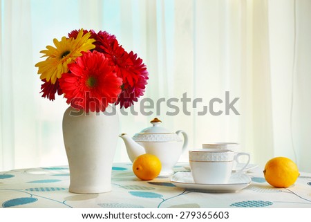 Colorful gerbera in vase with teapot, cups and lemons on table on curtains background