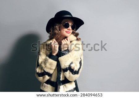 Portrait of beautiful model in fur coat, hat and sunglasses on gray background