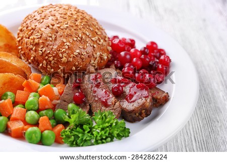 Beef with cranberry sauce, roasted potato slices, vegetables and bun on plate, on color wooden background