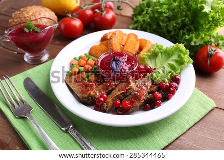 Beef with cranberry sauce, roasted potato slices, vegetables on plate, on  wooden background