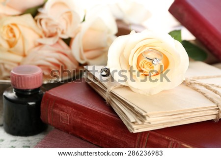 Wedding ring in fresh rose flower with old book and letters on color wooden table background. Vintage concept