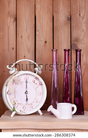 Interior design with alarm clock, decorative watering pot and vases on tabletop on wooden planks background