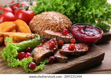 Beef with cranberry sauce, roasted potato slices and bun on cutting board, on wooden background