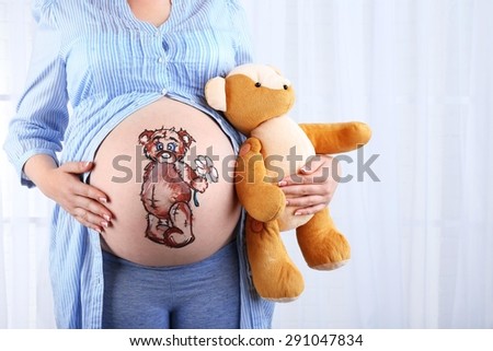Beautiful young pregnant with baby toy  and picture on her belly, on light background