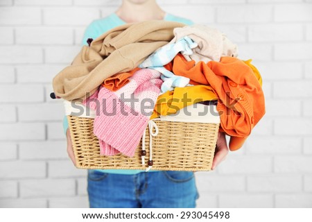 Woman holding basket with heap of different clothes, on bricks wall background
