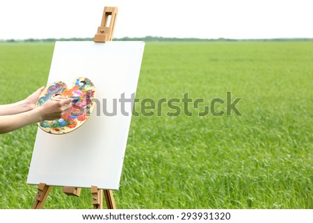 Female hand holding palette and easel with canvas in green field