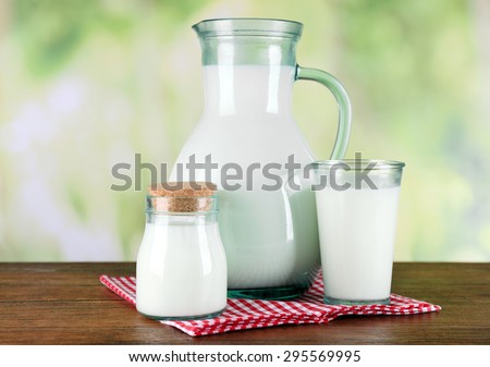 Pitcher, jar and glass of milk on wooden table, on nature background