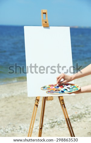Female hand holding palette with paints and easel with canvas on beach