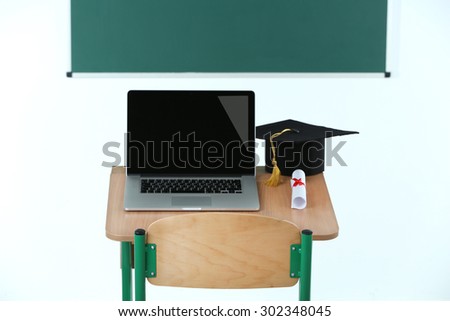 School desk with laptop, master hat and diploma in classroom