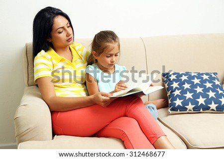 Beautiful little girl reading book with mother on sofa at home