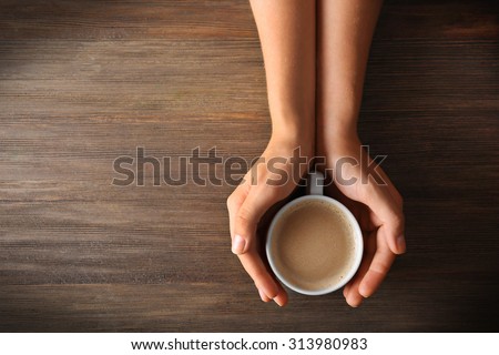 Female hands holding a cup of coffee with foam over wooden table, top view