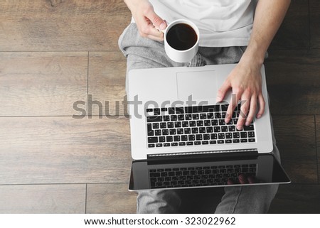 Young man sitting on floor with laptop and cup of coffee in room
