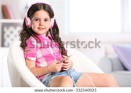 Attractive little girl sitting on comfortable chair and listening music with pink headphones in the room